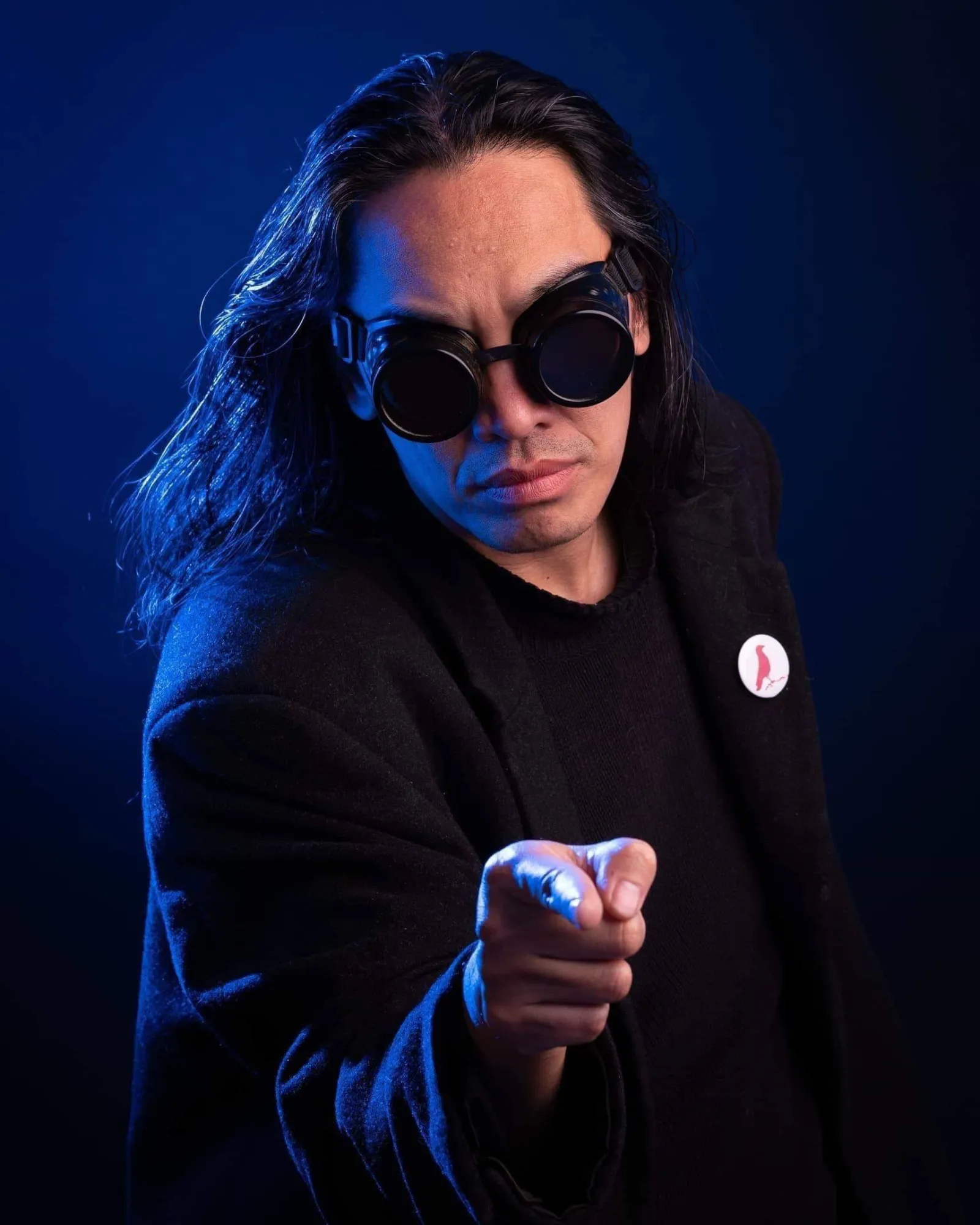 Bryan Thao Worra in round dark goggles and a black coat, pointing at the camera against a blue backdrop