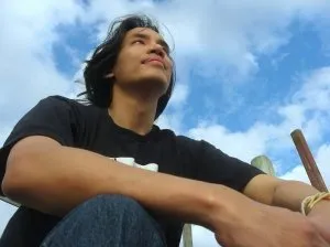 Man with windswept dark hair sitting outdoors under a blue, cloudy sky