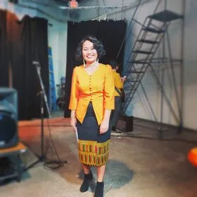 Kaysone Syonesa in an orange Lao blouse and woven sinh, standing on a stage